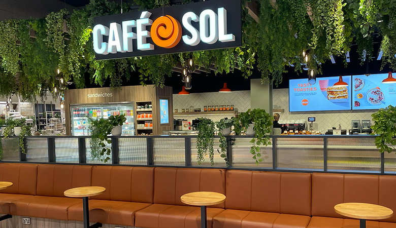 Interior view of a Cafe Sol restaurant location showing the illuminated brand sign, service counter with food displays, seating area with brown leather banquettes and round wooden tables, and menu boards